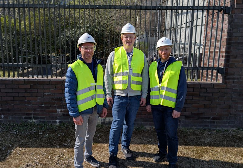 Drei Männer mit Schutzhelmen und gelben Warnwesten stehen im Freien vor einem Zaun und einer Backsteinmauer. Sie lächeln in die Kamera. Das Bild vermittelt den Kontext von GIS und Opensource, beispielsweise im Rahmen eines Projekts oder einer Besichtigung im technischen oder infrastrukturellen Bereich. OpenSource GIS.