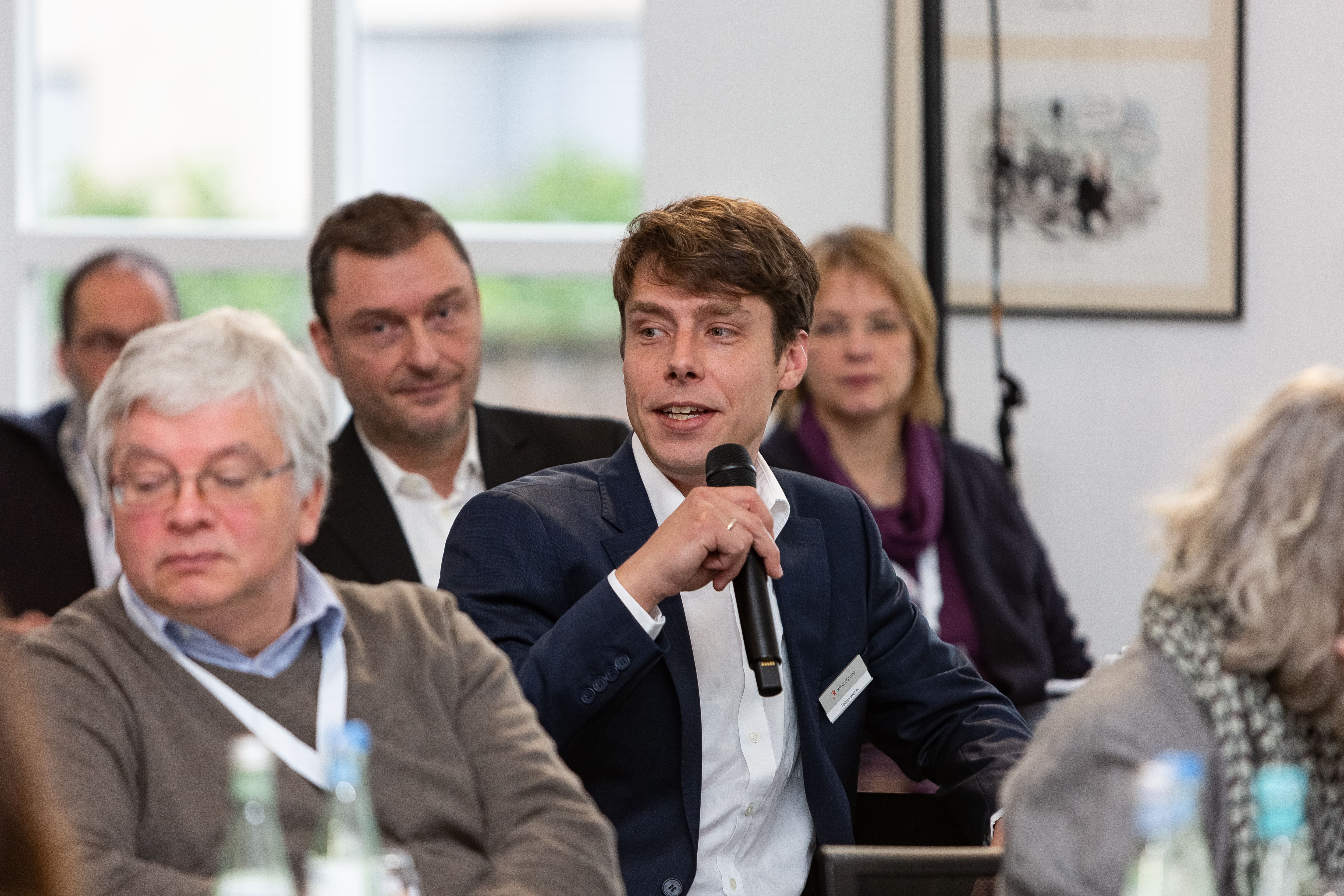 Ein Mann im Anzug spricht mit einem Mikrofon während der Where2B. Um ihn herum sitzen weitere Teilnehmende, die aufmerksam zuhören. Die Szene vermittelt den Austausch über Fachthemen wie GIS und Opensource.